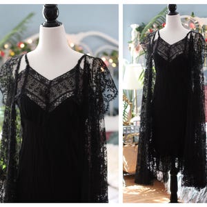 K&ouml;nnte beinhalten: Ein schwarzes Kleid mit V-Ausschnitt und zarten Spitzendetails. Das Kleid wird mit einem passenden Spitzenumhang kombiniert, wodurch eine elegante und gotische &Auml;sthetik entsteht. Das Kleid wird auf einer Schaufensterpuppe pr&auml;sentiert.