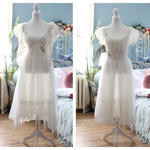 May include: Two white vintage dresses on display. The dresses feature delicate lace and floral embroidery. One dress has a longer, flowing skirt, while the other is knee-length. Both are displayed on white mannequins, showcasing their elegant design.