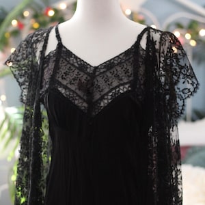 K&ouml;nnte beinhalten: Ein schwarzes Slip-Kleid mit V-Ausschnitt und zarten Spitzendetails. Das Kleid wird mit einem passenden Spitzen-Overlay kombiniert, wodurch ein geschichteter Effekt entsteht. Das Kleid wird auf einer Schaufensterpuppe pr&auml;sentiert.