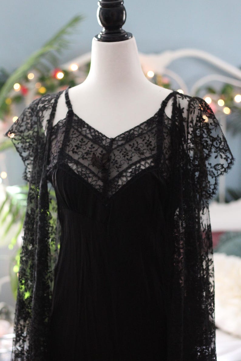 K&ouml;nnte beinhalten: Ein schwarzes Slip-Kleid mit V-Ausschnitt und zarten Spitzendetails. Das Kleid wird mit einem passenden Spitzen-Overlay kombiniert, wodurch ein geschichteter Effekt entsteht. Das Kleid wird auf einer Schaufensterpuppe pr&auml;sentiert.