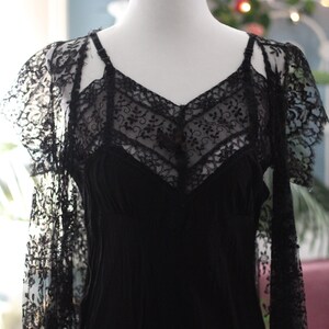 K&ouml;nnte beinhalten: Ein schwarzes Vintage-Kleid mit V-Ausschnitt und zarten Spitzendetails. Das Kleid hat Spaghettitr&auml;ger und wird auf einer Schaufensterpuppe pr&auml;sentiert. Die &Auml;rmel sind aus durchsichtiger Spitze gefertigt und verleihen dem Design einen Hauch von Eleganz.