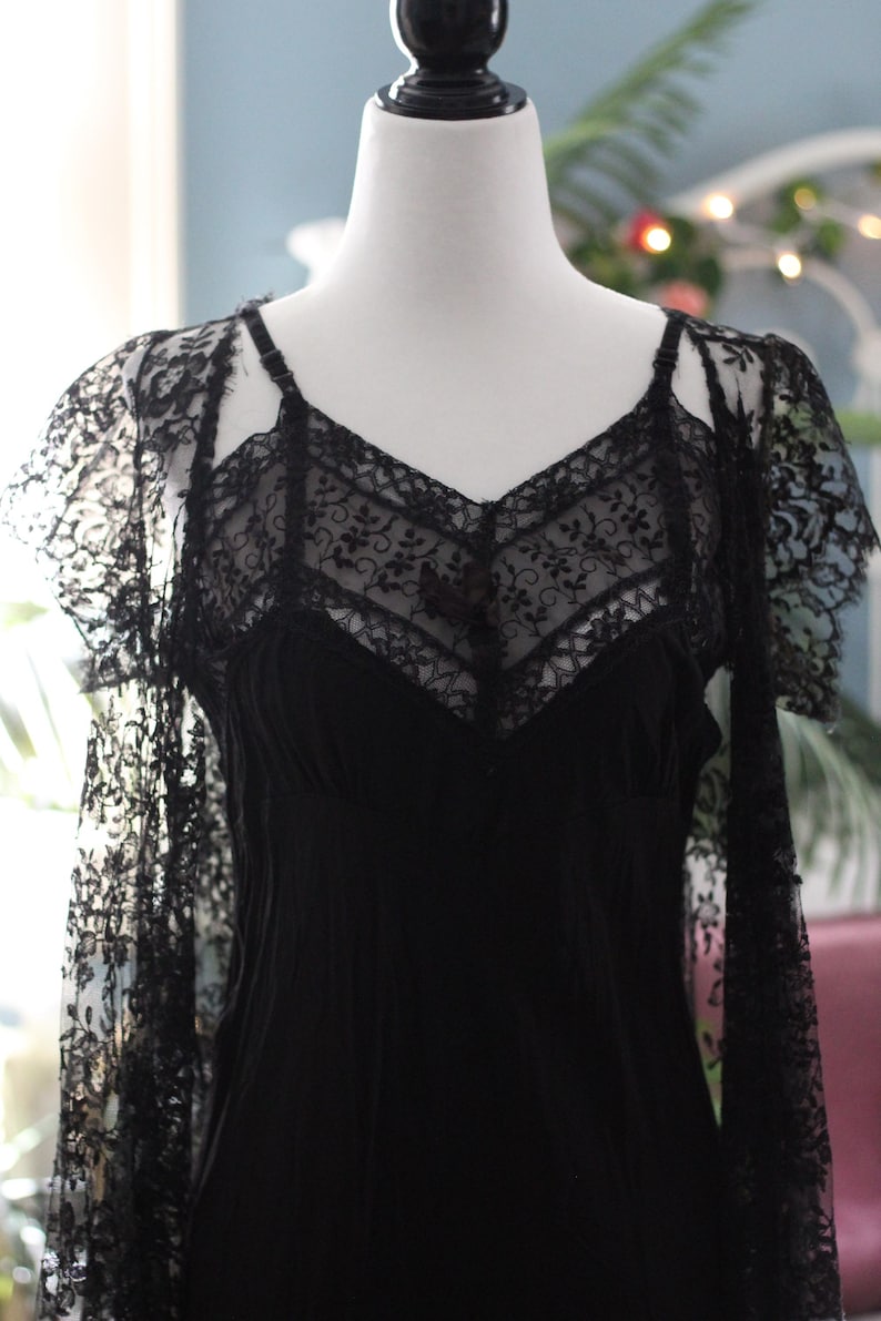K&ouml;nnte beinhalten: Ein schwarzes Vintage-Kleid mit V-Ausschnitt und zarten Spitzendetails. Das Kleid hat Spaghettitr&auml;ger und wird auf einer Schaufensterpuppe pr&auml;sentiert. Die &Auml;rmel sind aus durchsichtiger Spitze gefertigt und verleihen dem Design einen Hauch von Eleganz.