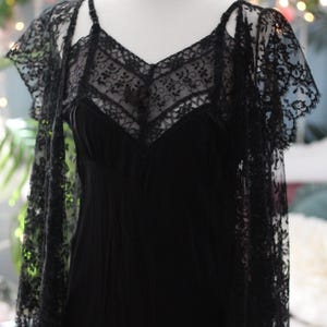 K&ouml;nnte beinhalten: Ein schwarzes Kleid im Vintage-Stil mit V-Ausschnitt und zarten Spitzendetails. Das Kleid wird mit einem passenden Spitzenschal kombiniert. Das Kleid wird auf einer Schaufensterpuppe pr&auml;sentiert.