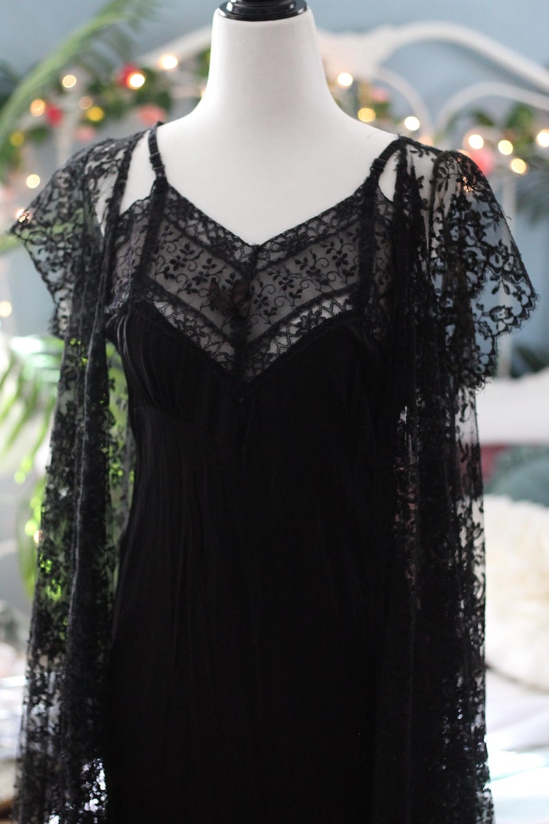 K&ouml;nnte beinhalten: Ein schwarzes Kleid im Vintage-Stil mit V-Ausschnitt und zarten Spitzendetails. Das Kleid wird mit einem passenden Spitzenschal kombiniert. Das Kleid wird auf einer Schaufensterpuppe pr&auml;sentiert.