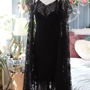 K&ouml;nnte beinhalten: Ein schwarzes Kleid im Vintage-Stil mit einer Spitzen-Overlay und passendem Spitzen-Cape. Das Kleid, das auf einer Schaufensterpuppe pr&auml;sentiert wird, hat einen V-Ausschnitt und Spaghettitr&auml;ger. Das Cape hat kurze &Auml;rmel und drapiert die Schultern.