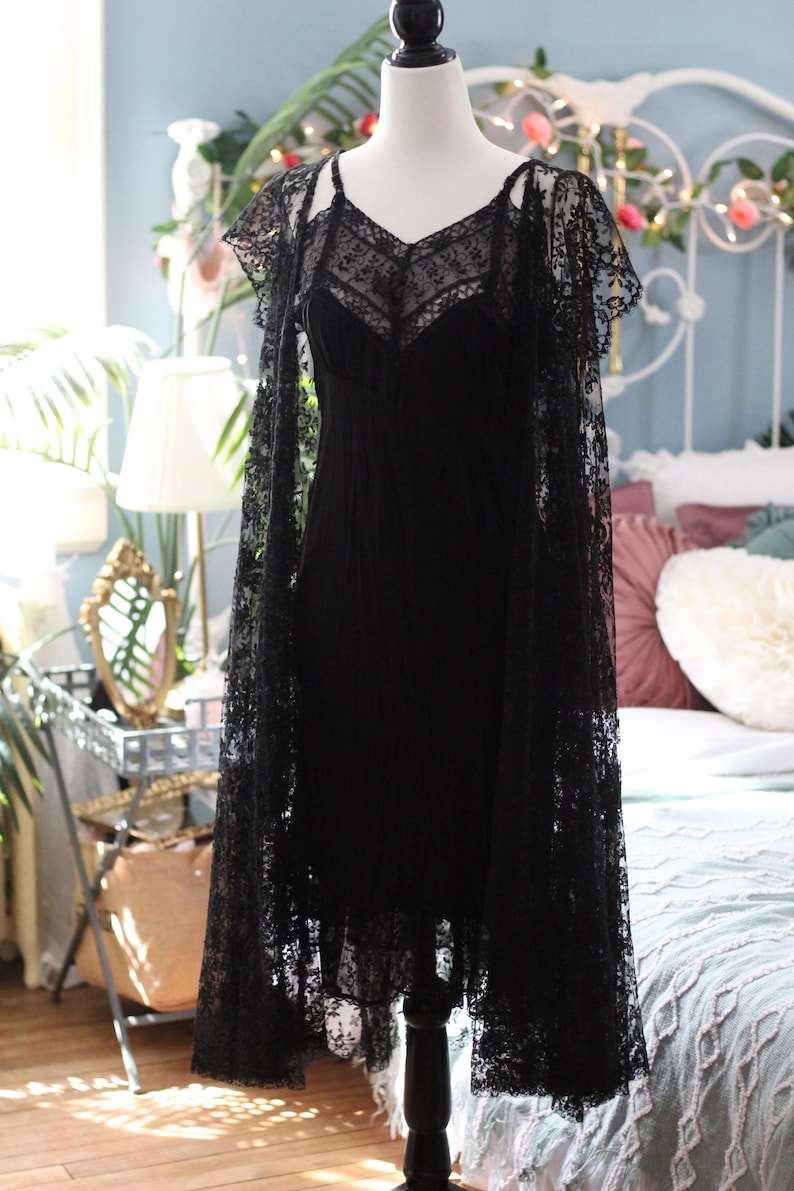 K&ouml;nnte beinhalten: Ein schwarzes Kleid im Vintage-Stil mit einer Spitzen-Overlay und passendem Spitzen-Cape. Das Kleid, das auf einer Schaufensterpuppe pr&auml;sentiert wird, hat einen V-Ausschnitt und Spaghettitr&auml;ger. Das Cape hat kurze &Auml;rmel und drapiert die Schultern.