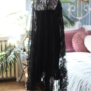 K&ouml;nnte beinhalten: Ein schwarzes Spitzenkleid auf einer Schaufensterpuppe. Das Kleid hat einen zarten Spitzeneinsatz, einen hohen Ausschnitt und einen flie&szlig;enden Rock. Die komplizierten Details der Spitze sind sichtbar und schaffen eine romantische und elegante &Auml;sthetik. Das Kleid wird auf einem schwarzen St&auml;nder ausgestellt.