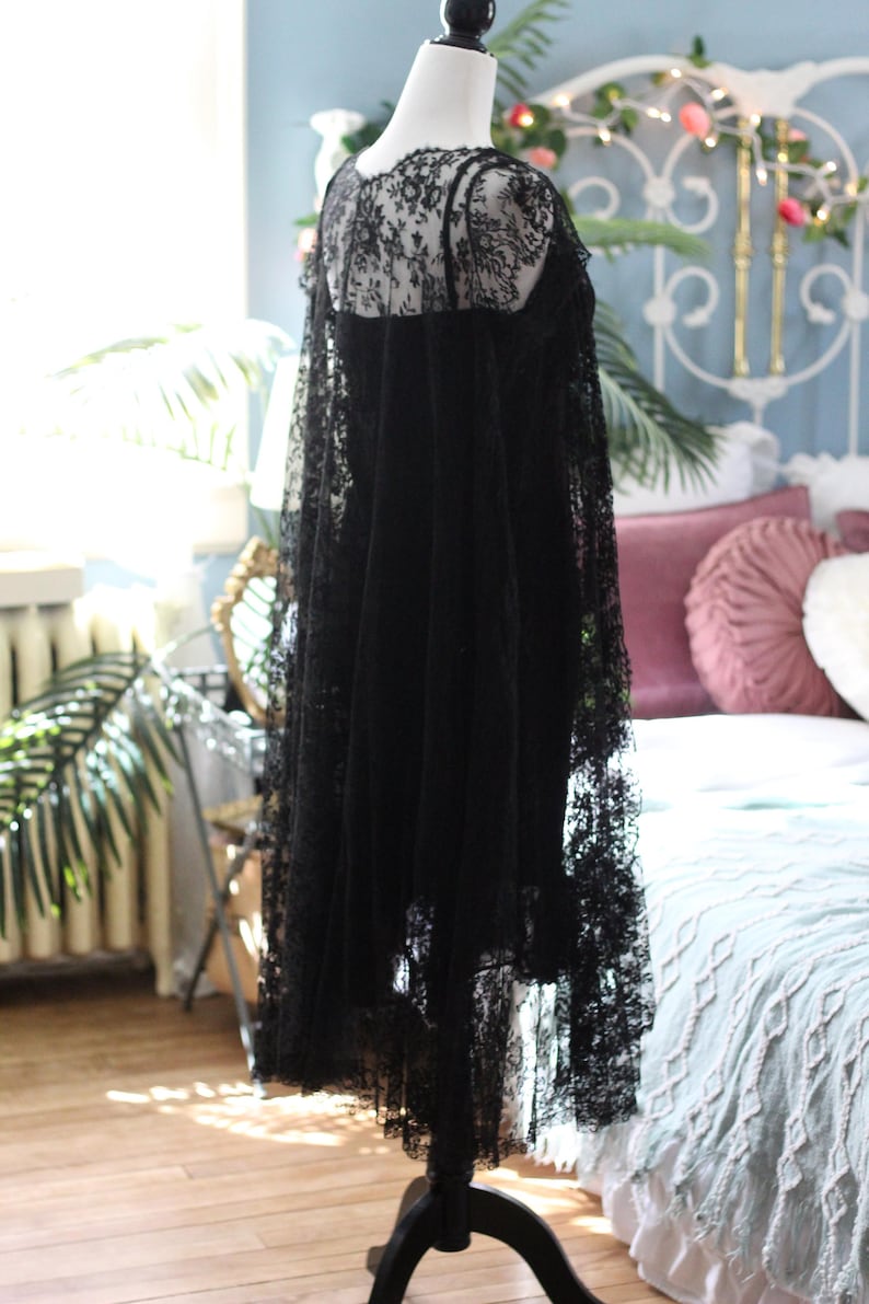 K&ouml;nnte beinhalten: Ein schwarzes Spitzenkleid auf einer Schaufensterpuppe. Das Kleid hat einen zarten Spitzeneinsatz, einen hohen Ausschnitt und einen flie&szlig;enden Rock. Die komplizierten Details der Spitze sind sichtbar und schaffen eine romantische und elegante &Auml;sthetik. Das Kleid wird auf einem schwarzen St&auml;nder ausgestellt.