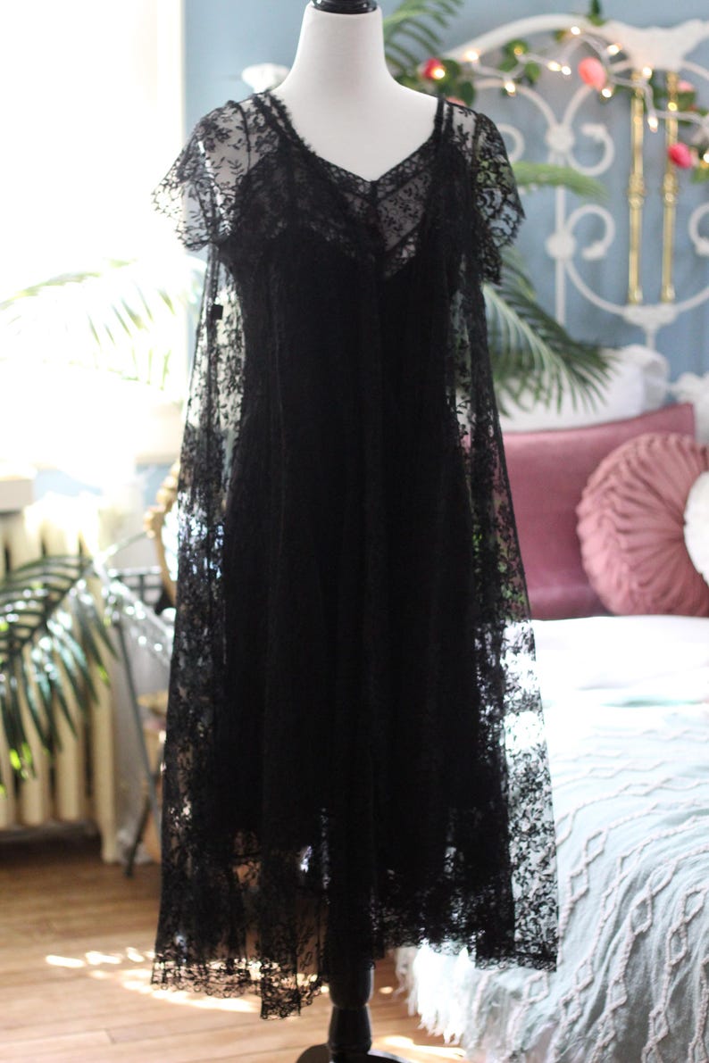 K&ouml;nnte beinhalten: Schwarzes Spitzenkleid auf einer Schaufensterpuppe. Das Kleid hat einen V-Ausschnitt, kurze &Auml;rmel und einen flie&szlig;enden Rock. Die filigrane Spitze bedeckt das gesamte Kleid und erzeugt ein zartes und elegantes Aussehen. Das Kleid ist bodenlang.