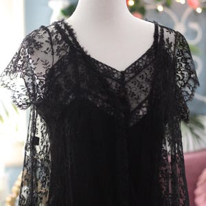 K&ouml;nnte beinhalten: Schwarzes Spitzenkleid mit V-Ausschnitt und kurzen &Auml;rmeln, pr&auml;sentiert auf einer Schaufensterpuppe. Das Kleid weist aufwendige florale Spitzendetails auf, die ein zartes und elegantes Aussehen erzeugen. Das Kleid ist ein klassisches St&uuml;ck.