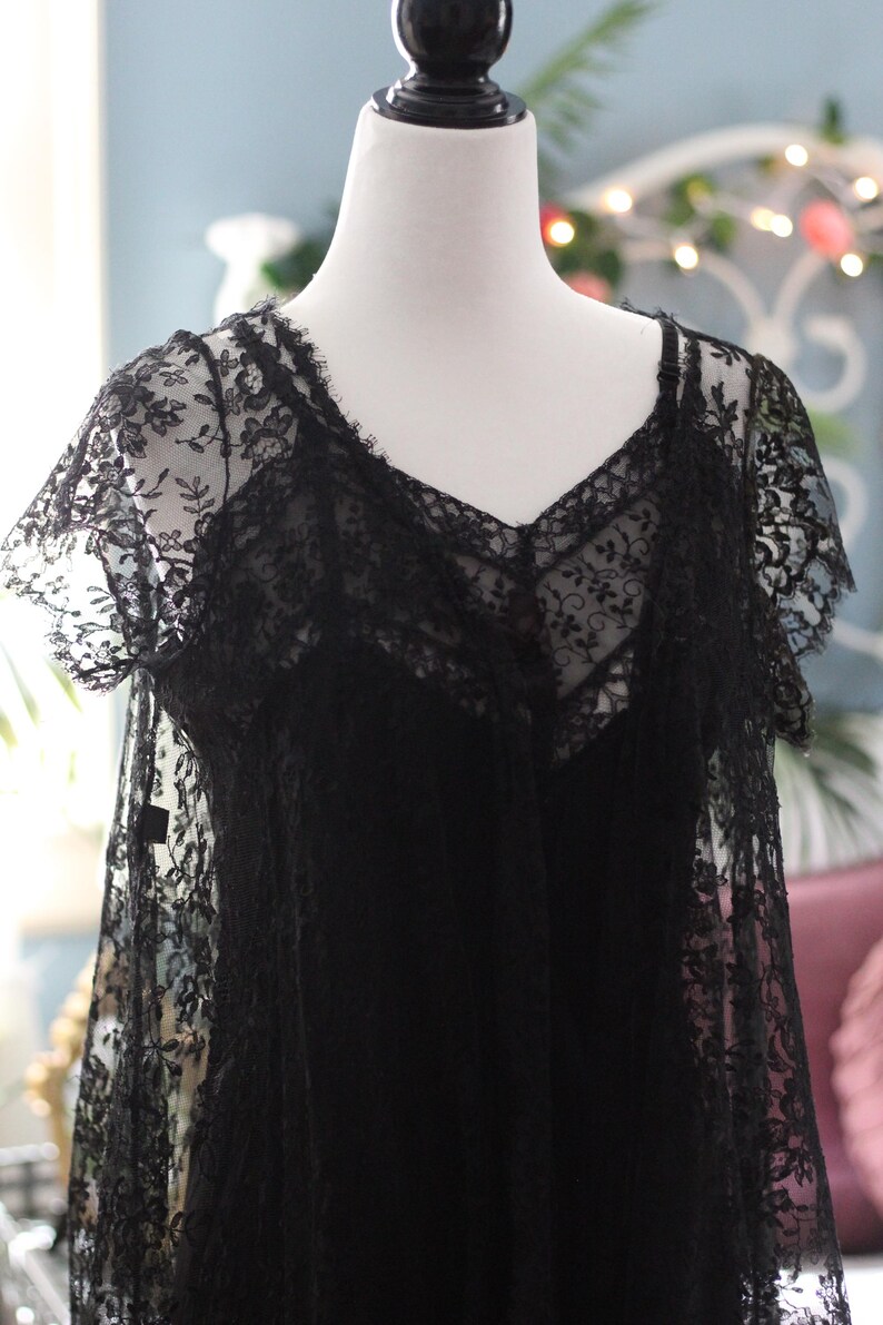 K&ouml;nnte beinhalten: Schwarzes Spitzenkleid mit V-Ausschnitt und kurzen &Auml;rmeln, pr&auml;sentiert auf einer Schaufensterpuppe. Das Kleid weist aufwendige florale Spitzendetails auf, die ein zartes und elegantes Aussehen erzeugen. Das Kleid ist ein klassisches St&uuml;ck.