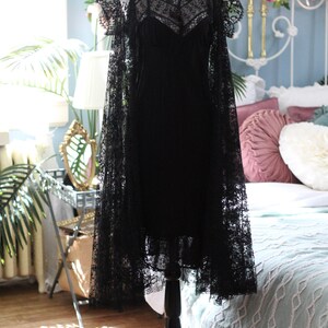 K&ouml;nnte beinhalten: Ein schwarzes Kleid im Vintage-Stil und ein Spitzenumhang auf einer Schaufensterpuppe. Das Kleid hat einen V-Ausschnitt und Spaghettitr&auml;ger mit zarter Spitzenauflage. Der Umhang hat kurze &Auml;rmel und aufwendige Spitzendetails.