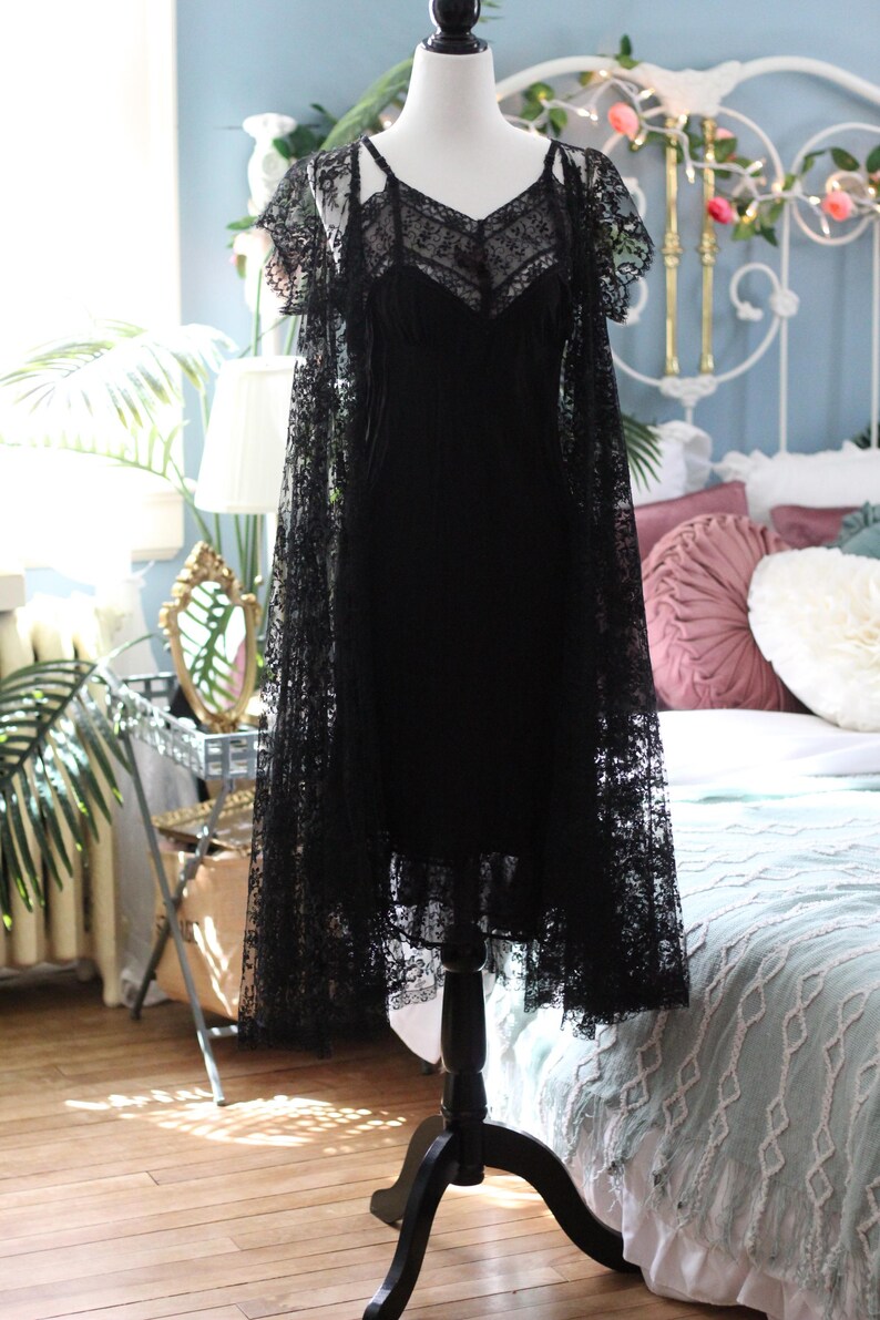 K&ouml;nnte beinhalten: Ein schwarzes Kleid im Vintage-Stil und ein Spitzenumhang auf einer Schaufensterpuppe. Das Kleid hat einen V-Ausschnitt und Spaghettitr&auml;ger mit zarter Spitzenauflage. Der Umhang hat kurze &Auml;rmel und aufwendige Spitzendetails.