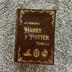 Może przedstawiać: Brązowa książka w skórzanej oprawie z pozłacanymi napisami i ozdobnymi akcentami. Tytuł to "J.K. Rowling's Harry Potter Years 1-3".