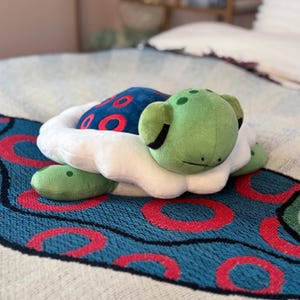 May include: A plush turtle toy with a green head and feet, a blue shell with red circles, and a white ruffled edge. The toy is resting on a blue patterned blanket with red circles.