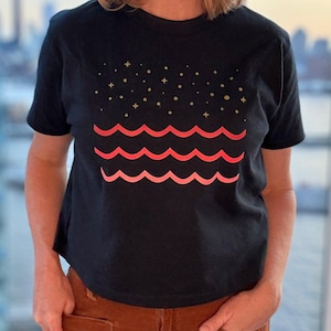 Op de afbeelding: Een zwart T-shirt met korte mouwen voorzien van een grafische print van drie horizontale golvende lijnen in roze en koraaltinten, geplaatst onder een patroon van kleine goudkleurige sterretjes en stippen op de borst.