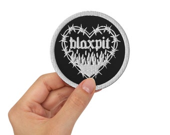 Peut inclure: Un écusson rond noir avec un motif brodé blanc. Le motif présente le mot "bloxpit" dans une police gothique, entouré d'un motif de fil de fer barbelé en forme de cœur. L'écusson est tenu dans une main.