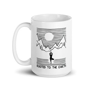May include: A white ceramic mug featuring a black line art illustration of a person in a yoga tree pose centered below a mountain range and a sun. The text "ROOTED TO THE EARTH" is printed in black capital letters at the bottom of the design.