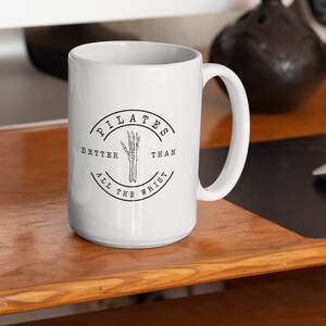 May include: White ceramic mug with a black and white graphic of a skeleton hand and the text "Pilates Better Than All The Wrist".