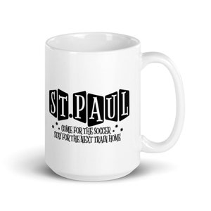 May include: White ceramic mug with a black graphic that reads "ST. PAUL" in a blocky font. The graphic also includes the text "COME FOR THE SOCCER. STAY FOR THE NEXT TRAIN HOME."