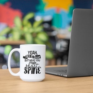 May include: A white ceramic mug with a black handle. The mug features the text "Yeah, Sex is Good But Have You Tried Short Spine" in black lettering. A laptop is visible in the background.