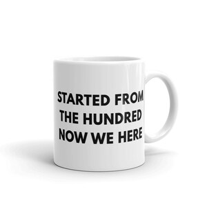 May include: White ceramic mug with the text "STARTED FROM THE HUNDRED NOW WE HERE" in black, a humorous statement. The mug has a curved handle and is set against a white background.