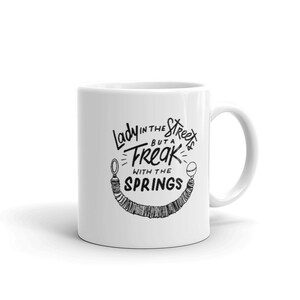 May include: White ceramic mug with a black graphic design. The text on the mug reads "Lady in the Streets but a Freak with the Springs". The design includes a stylized illustration of springs. The mug has a curved handle.
