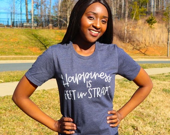 May include: A woman wearing a dark blue t-shirt with the text "Happiness is feet in straps" in white lettering.