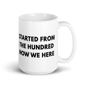 May include: A white ceramic mug featuring the black printed text STARTED FROM THE HUNDRED NOW WE HERE. The mug has a cylindrical body, a smooth finish, and a curved handle on the right side, providing a clean and simple design for daily beverage use.