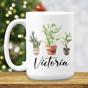 May include: White ceramic mug with a black handle. The mug has a watercolor illustration of three potted succulents. The text "Victoria" is printed in black below the succulents.