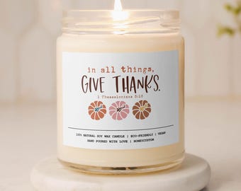 May include: A white candle with a clear glass jar and a label that reads "in all things, GIVE THANKS. 1 Thessalonians 5:18". The label also includes three stylized pumpkins in orange, pink, and brown. The candle is on a white pedestal.