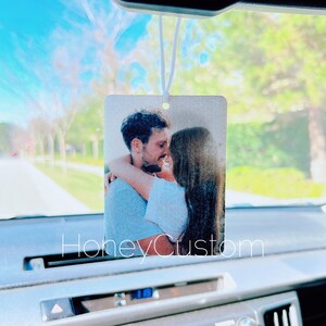 May include: A white car air freshener with a photo of a couple hugging. The photo is printed on a white background and the air freshener is hanging from the rearview mirror.