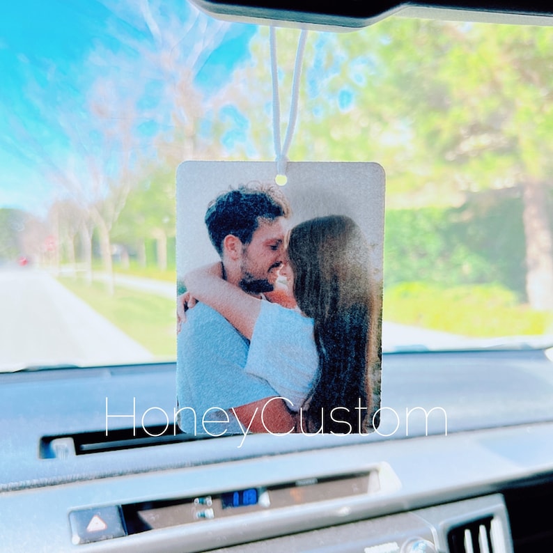 May include: A white car air freshener with a photo of a couple hugging. The photo is printed on a white background and the air freshener is hanging from the rearview mirror.