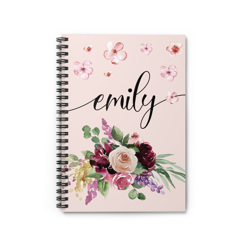May include: A pink spiral notebook with the name "emily" written in black cursive script. The cover features a watercolor floral design with burgundy, pink, and white flowers, green leaves, and a scattering of pink flowers.