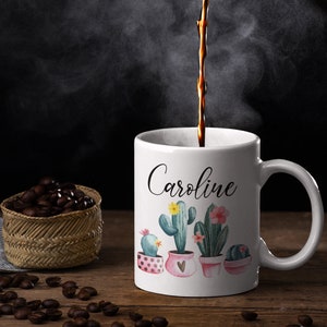 May include: White ceramic coffee mug with a black cursive name "Caroline" and a watercolor design of cacti in pink and green pots.