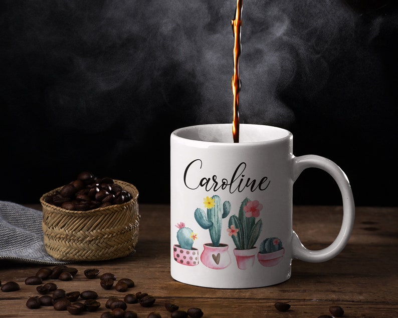 May include: White ceramic coffee mug with a black cursive name "Caroline" and a watercolor design of cacti in pink and green pots.
