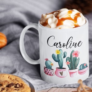 May include: White ceramic mug with a black cursive name "Caroline" and a design of four potted cacti with pink, yellow, and green flowers. The mug is filled with a creamy beverage topped with caramel sauce.