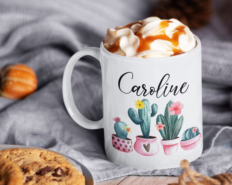 May include: White ceramic mug with a black cursive name "Caroline" and a design of four potted cacti with pink, yellow, and green flowers. The mug is filled with a creamy beverage topped with caramel sauce.