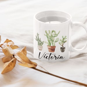 May include: White ceramic mug with a watercolor design of three potted cacti and the name "Victoria" written in black cursive.