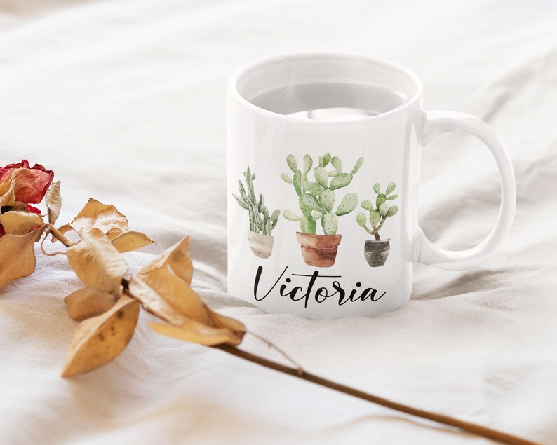 May include: White ceramic mug with a watercolor design of three potted cacti and the name "Victoria" written in black cursive.