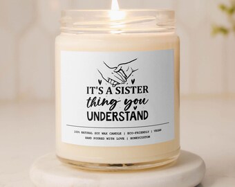 May include: A lit soy wax candle in a glass jar with a white label. The label reads "IT'S A SISTER thing you UNDERSTAND" with a graphic of two hands holding each other. The candle sits on a white, round coaster.