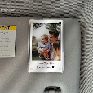 May include: A clear acrylic car visor photo holder with a family photo. The photo shows a father holding a baby, with a woman standing beside them. The text "Drive Safe, Dad. We Love You!" is printed below the photo.
