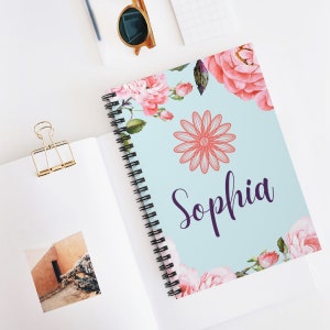 May include: A light blue spiral notebook with a floral design and the name "Sophia" printed in purple. The notebook features pink roses, green leaves, and a large pink flower in the center.