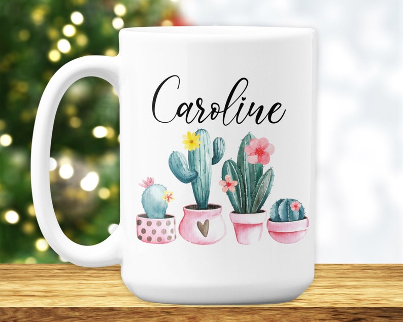 May include: White ceramic mug with a black cursive name "Caroline" and a watercolor illustration of five potted cacti with pink and yellow flowers.