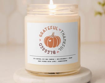 May include: A white candle with a glass jar and a label that says "Grateful Blessed Thankful" with a pumpkin graphic. The candle is lit and the wax is melted.