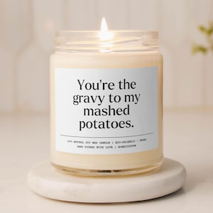 May include: A white candle with a clear glass jar and a black label that reads "You're the gravy to my mashed potatoes." The candle is lit and sitting on a white surface.