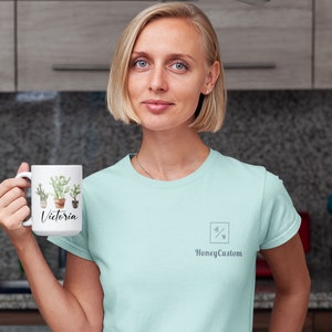 May include: A white ceramic mug with a cactus design and the name "Victoria" printed on it. The mug is being held by a person wearing a light blue t-shirt with a small logo that says "HoneyCustom".