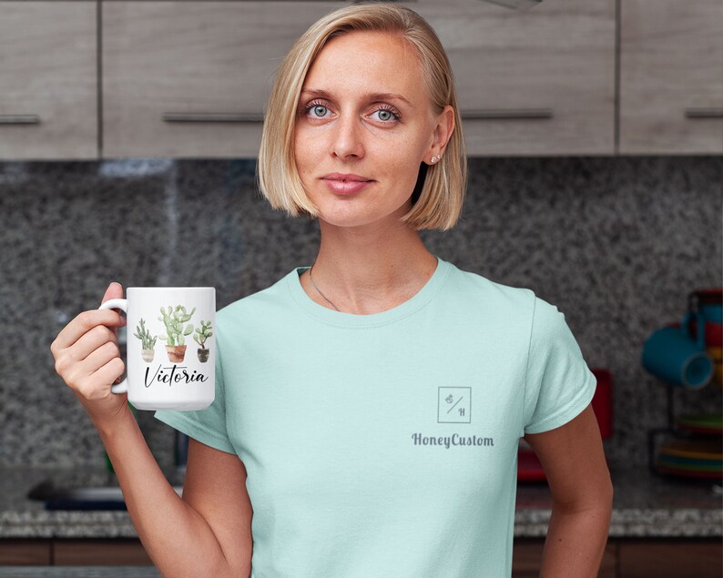 May include: A white ceramic mug with a cactus design and the name "Victoria" printed on it. The mug is being held by a person wearing a light blue t-shirt with a small logo that says "HoneyCustom".