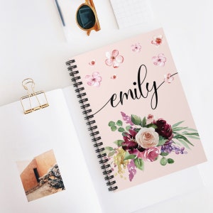 May include: A pink spiral notebook with the name "emily" written in black cursive. The cover features a watercolor floral design with pink, purple, and green flowers.