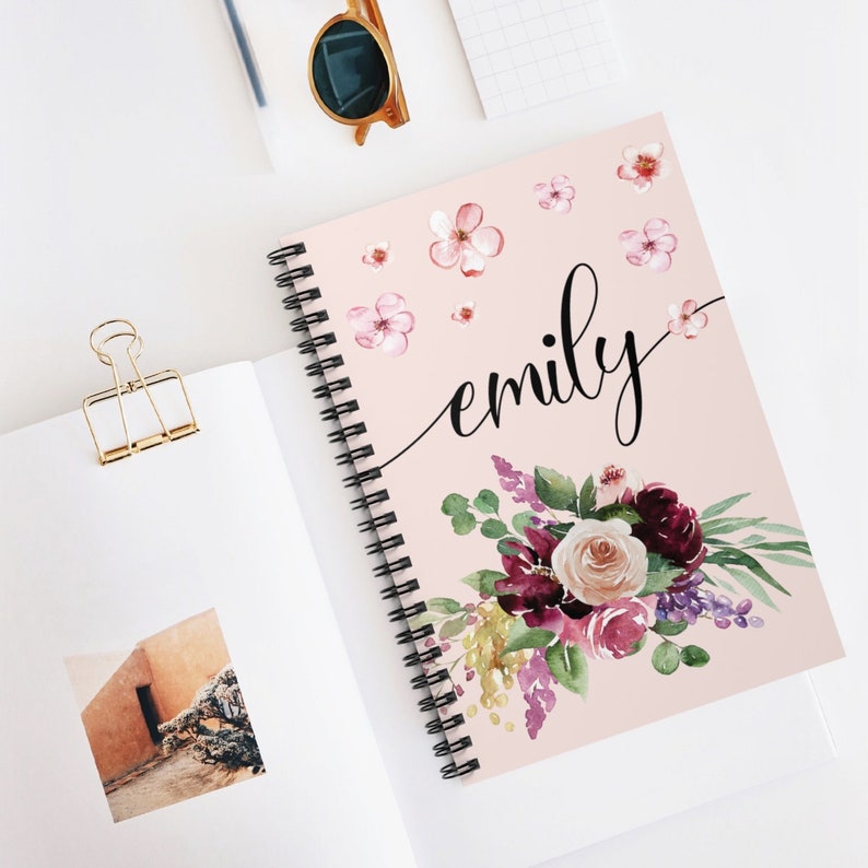 May include: A pink spiral notebook with the name "emily" written in black cursive. The cover features a watercolor floral design with pink, purple, and green flowers.